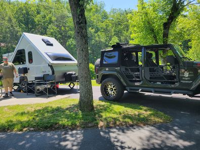 A jeep parked next to a trailer in the woods.