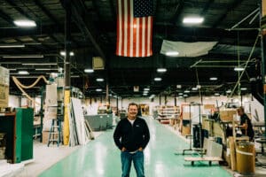 A man standing in an industrial warehouse with american flag overhead.