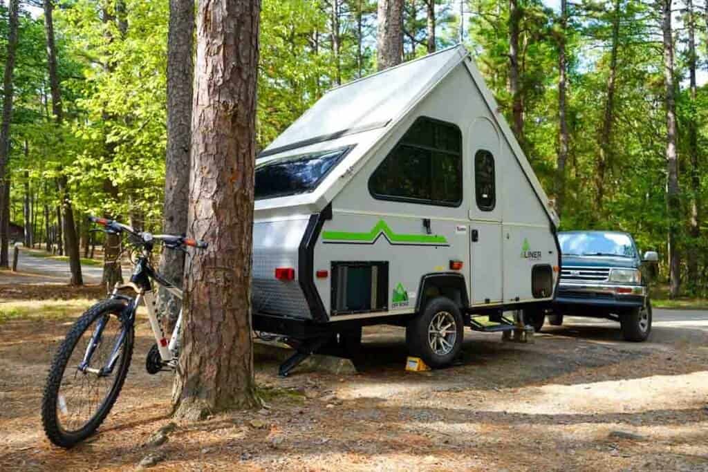 A travel trailer parked in a forested campground with a bicycle leaning against a tree in the foreground.